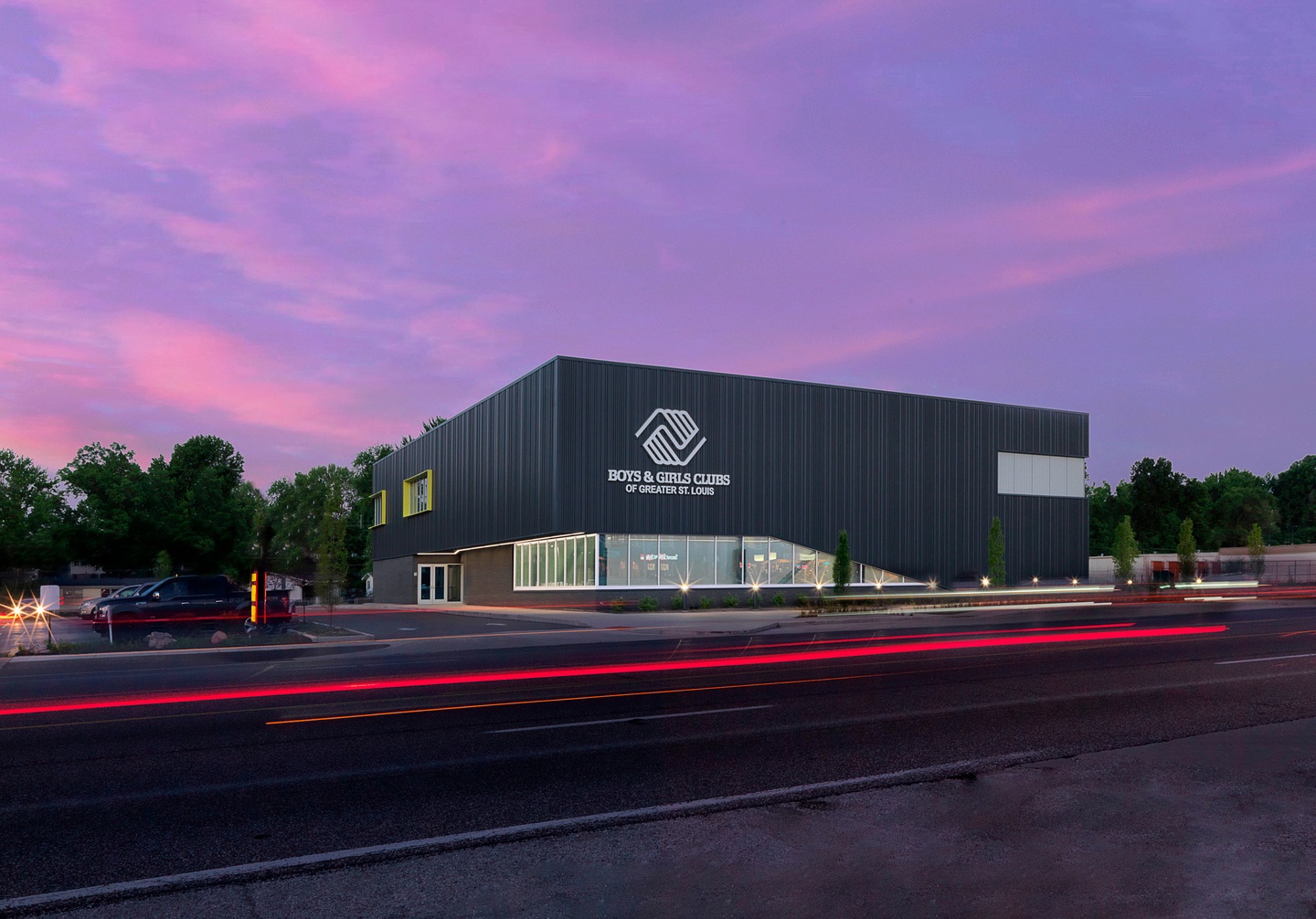 Photo of the exterior facade of new Boys & Girls club building, with a contemporary design including a bank of floor-to-ceiling first floor windows wrapping around the front corner of the building. The photo is shot at dusk with a purple sky and the street in front in view.