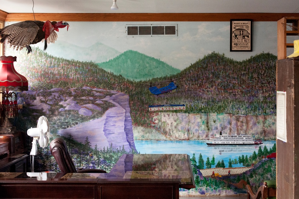 Office with a marble-topped desk, leather office chair, fan, fringed lamp, and taxidermied turkey. The back wall is painted with a mural of midwest landscape. A riverboat floats down the river and a military helicopter flies overhead with a banner advertising "Big Bags USA."