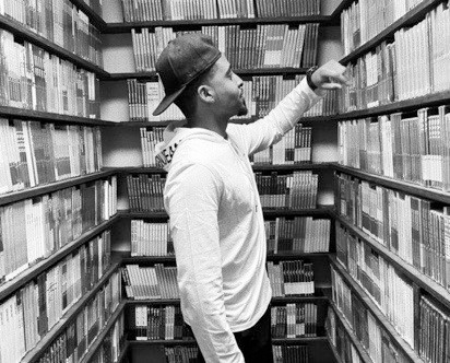 A Dominican American man stands in profile in an aisle of shelves lined with books. He wears a backwards baseball cap and reaches up toward a book on one of the shelves.
