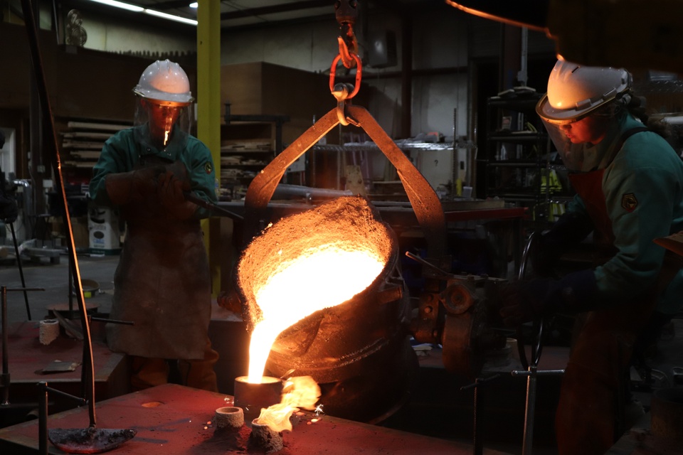 Two people in PPE look on as red hot melted steel is poured into a mold