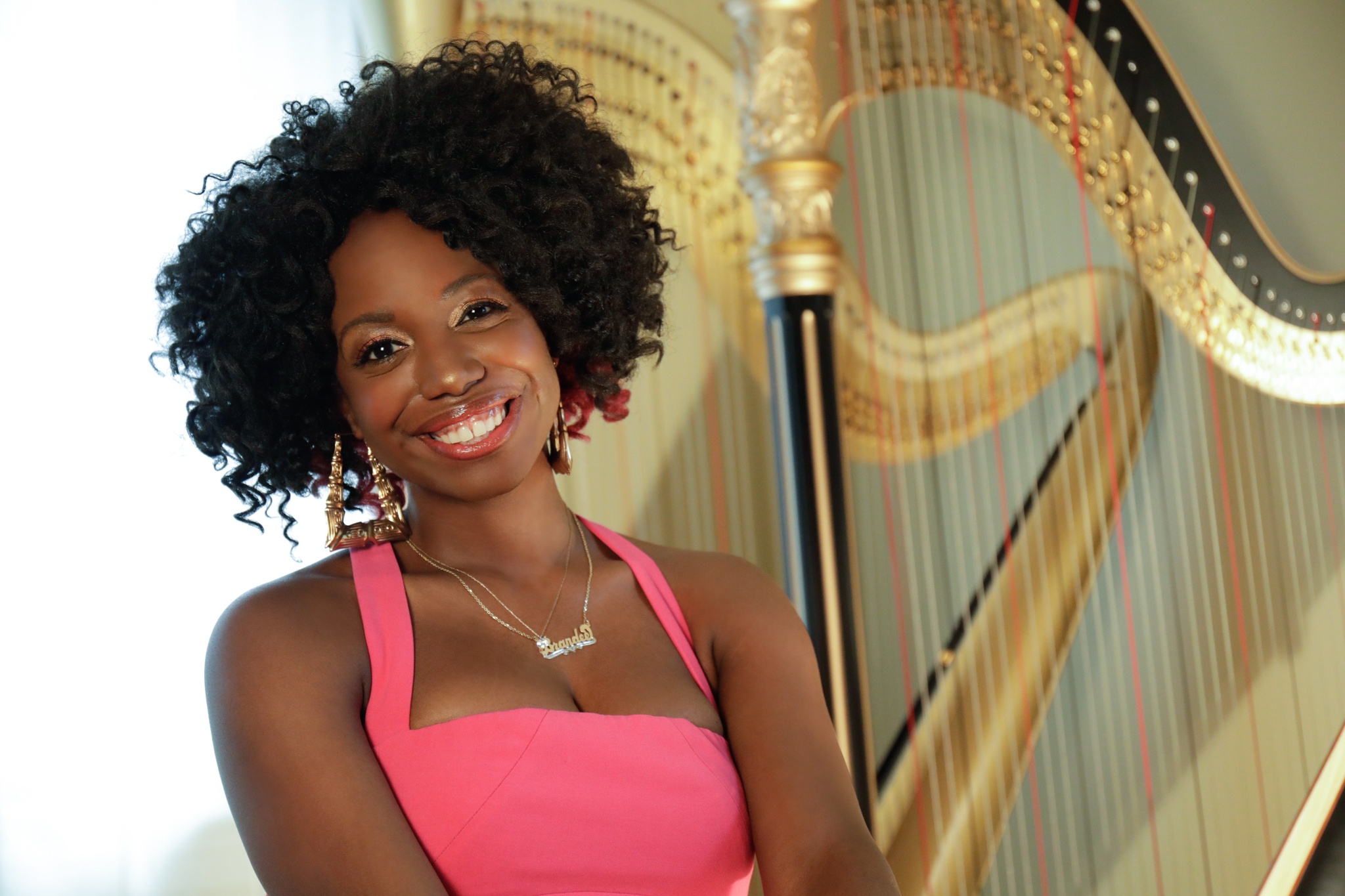 A Black woman in a pink dress with shoulder straps stands with two gilded harps behind her.