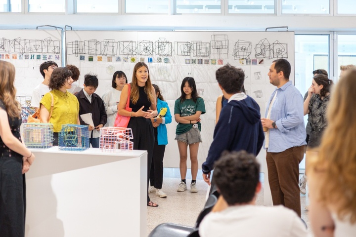 Students stand around a professor at an exhibition of their architectural models.