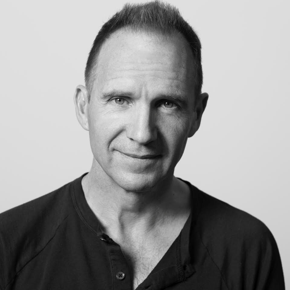 A black-and-white portrait of actor Ralph Fiennes, a white man with short brown hair. He smiles softly and wears a dark henley t-shirt.