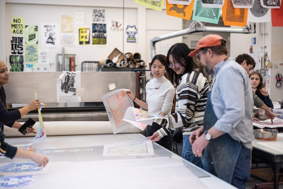 Three people hold and look at layers of painted and printed media in a workshop decorated with colorful prints.