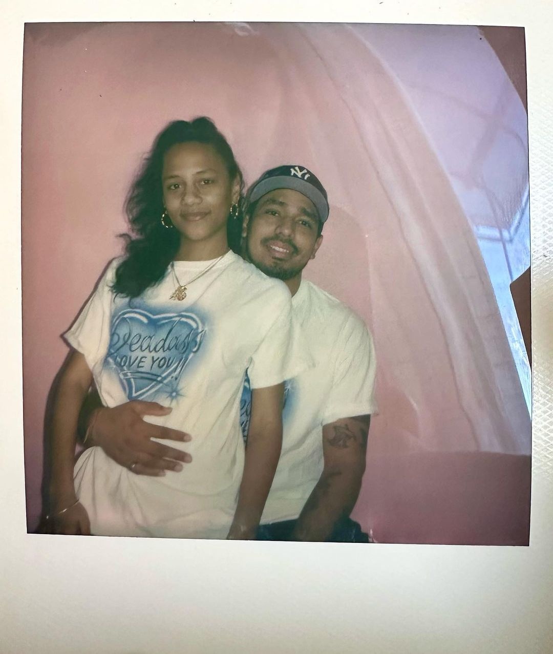 A Polaroid photo of two Latinx people wearing white tshirts. A woman sits in a man's lap. The man has his hand around her waist and looks out over her shoulder.