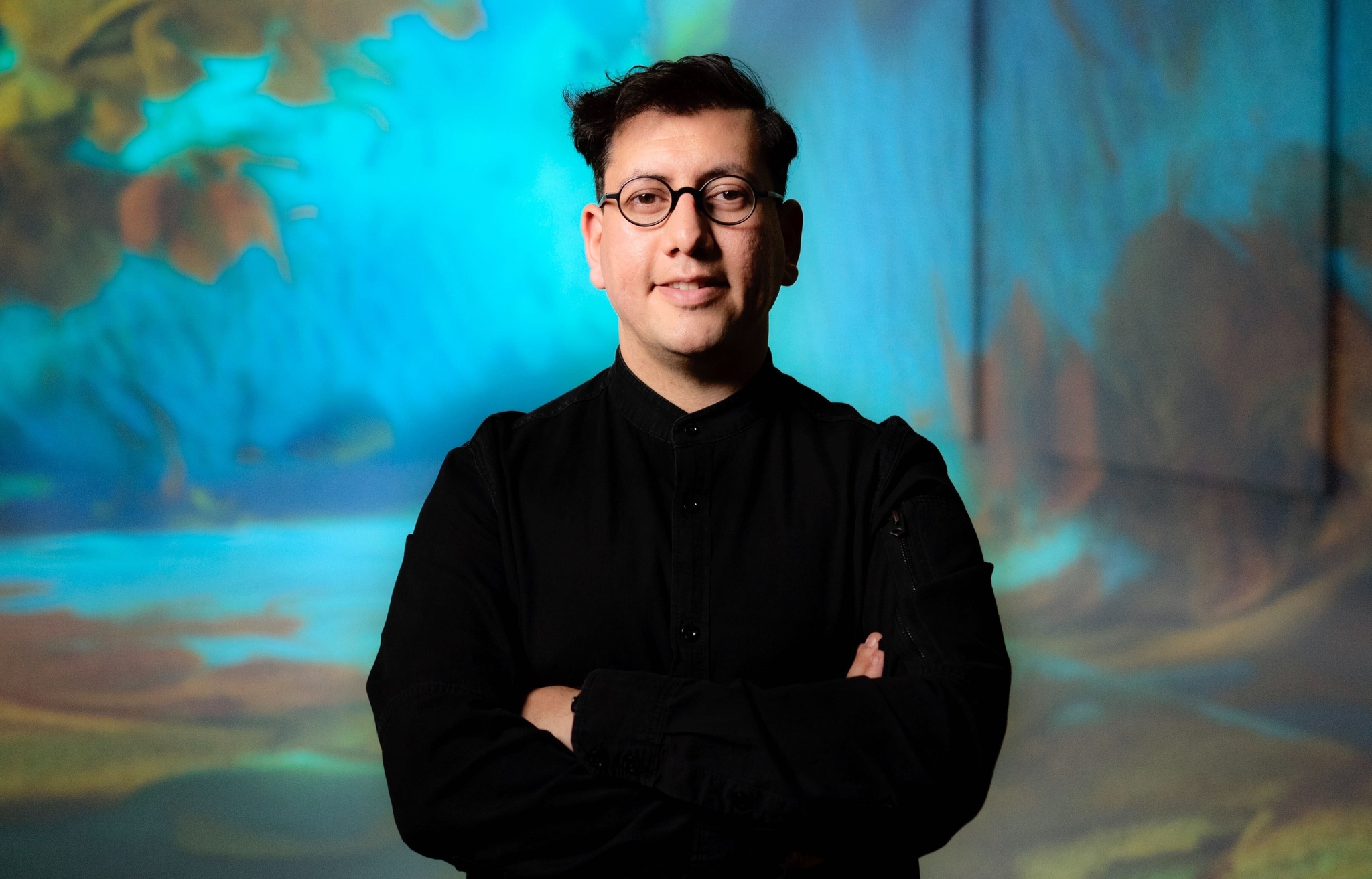 The artist Refik Anadol standing with arms crossed, wearing a black shirt and black-framed glasses, against a background with an abstract image recalling a forest or mountainous landscape 