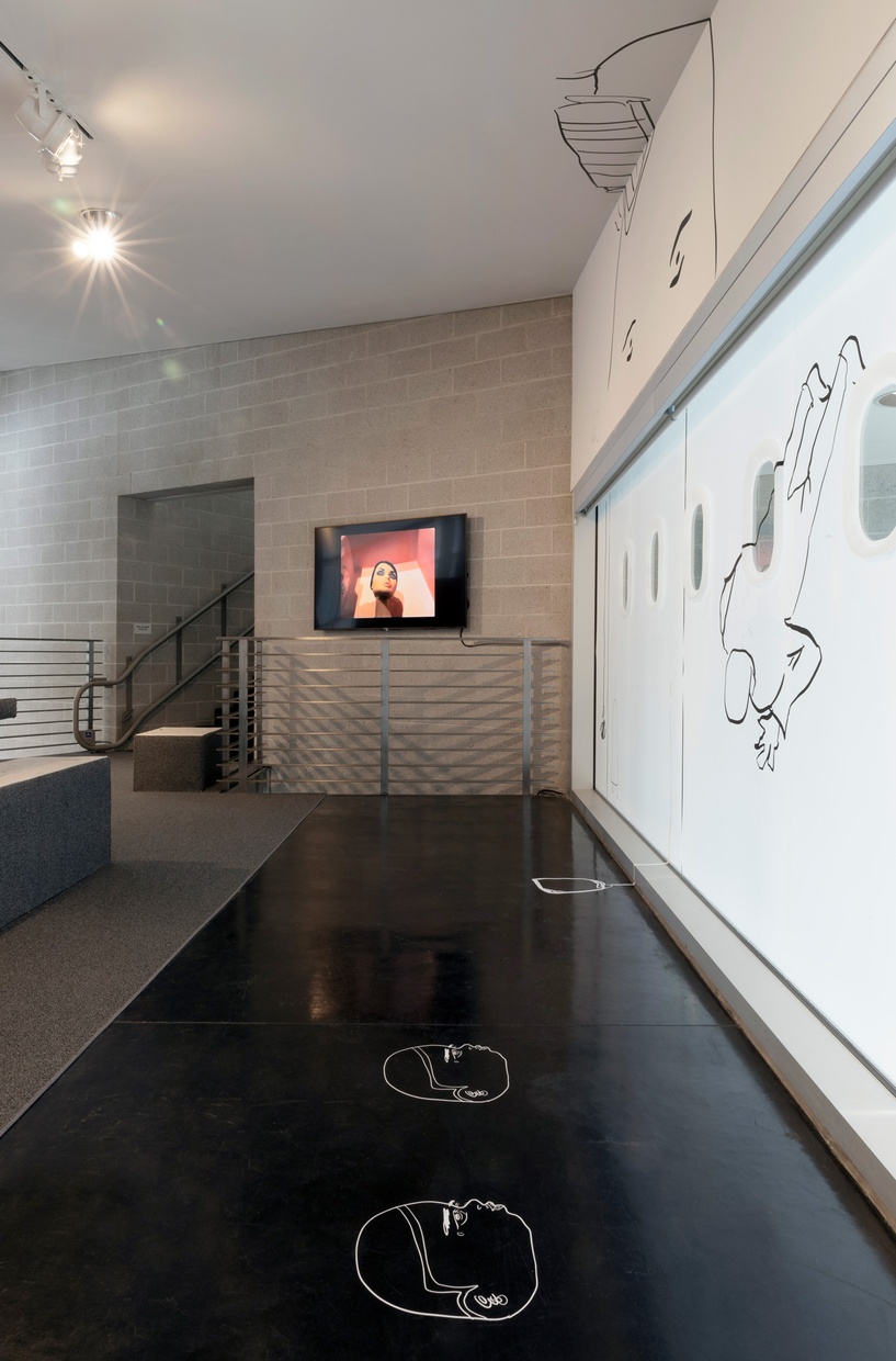 A room features white walls and a window on the right wall with black line drawings, and a gray brick wall to the left with a video animation playing on a television.