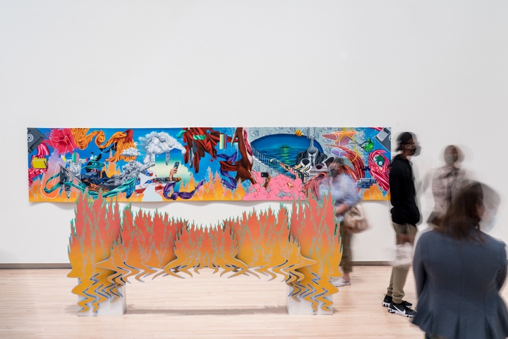 Long, brightly colored painting of highly rendered objects such as fabric, clouds, astronauts, hands holding up smartphones, a staircase, and headless bodies in suits floating in an abstract space. In front of the painting is installed a wooden, painted stage prop that looks like flames. People walk between the painting and the prop.