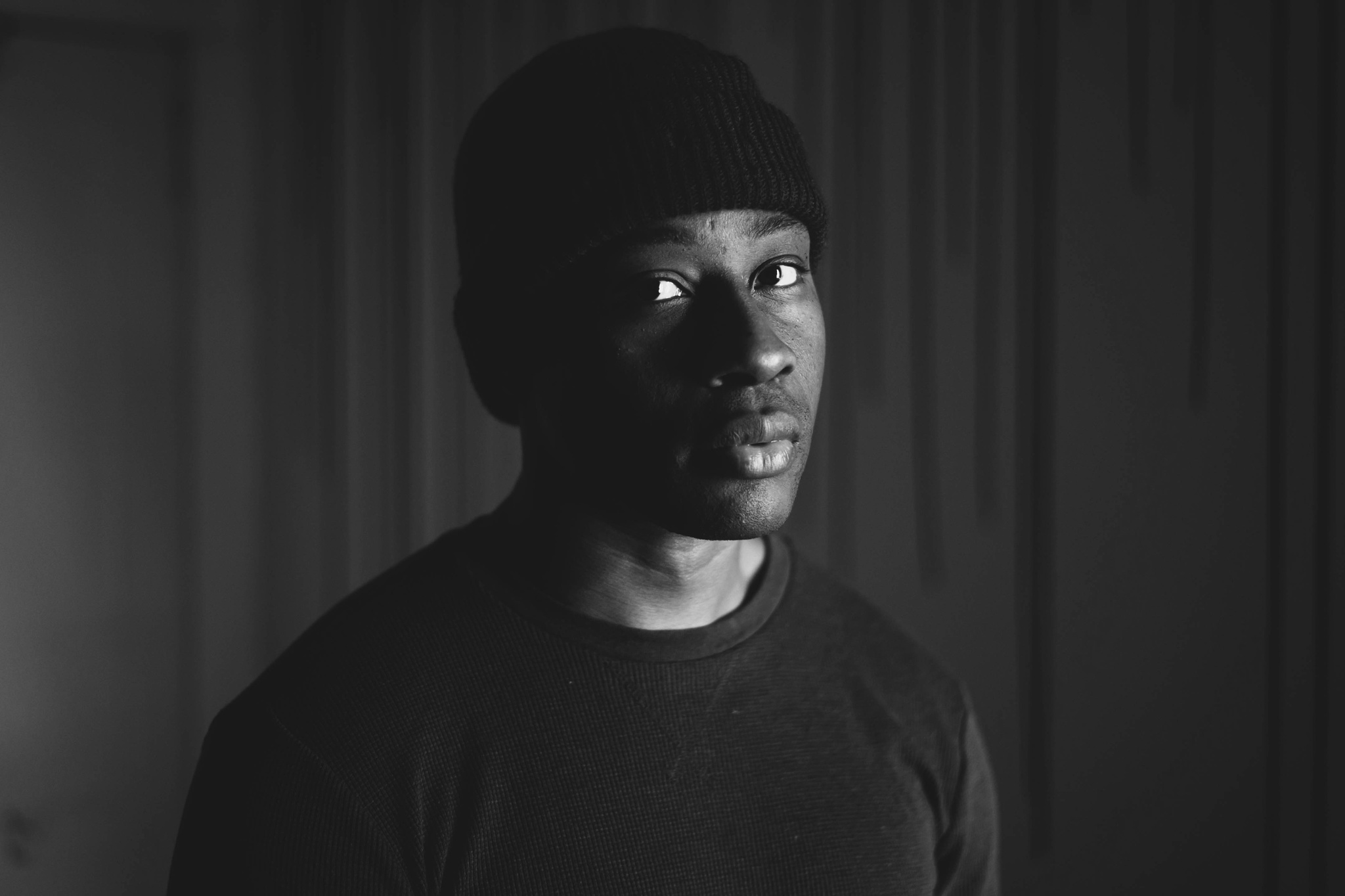 A black-and-white photo of Elliott Ashby, who wears a dark crewneck shirt and beanie. Light falls on the left side of Ashby's face, leaving the right side in dramatic shadow. 