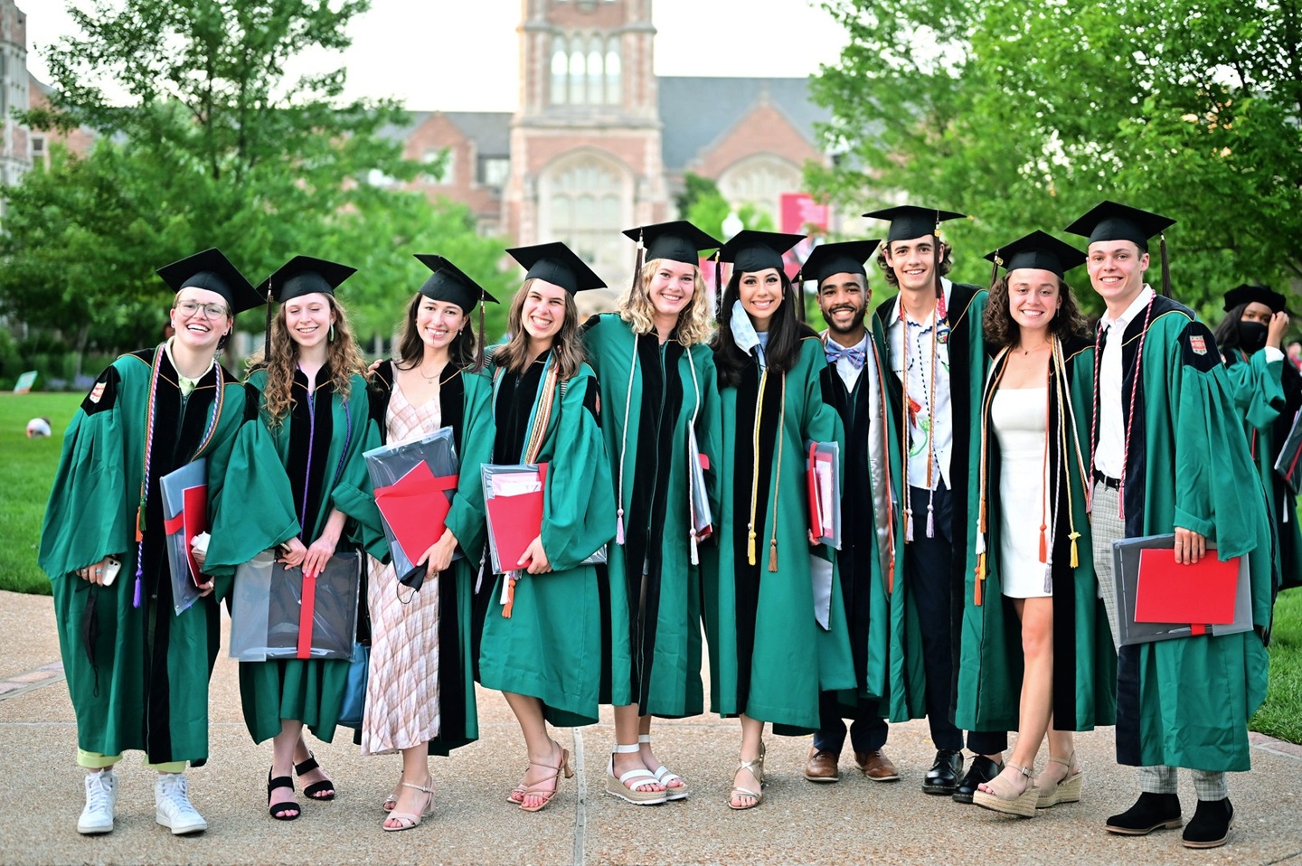 A group of students in green commencement gowns and black mortar boards poses together, with a Collegiate Gothic style building and leafy green trees in the background.