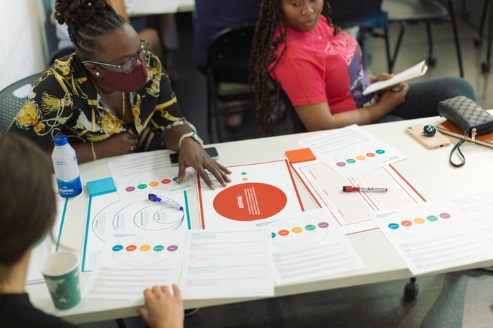 Three individuals actively engage with a tabletop filled with papers including graphs, diagrams, and text.