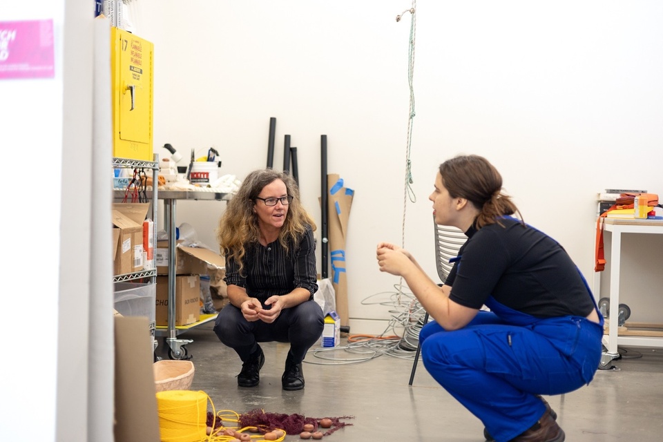 Two artists crouch to look at in-progress work on the floor of a studio.