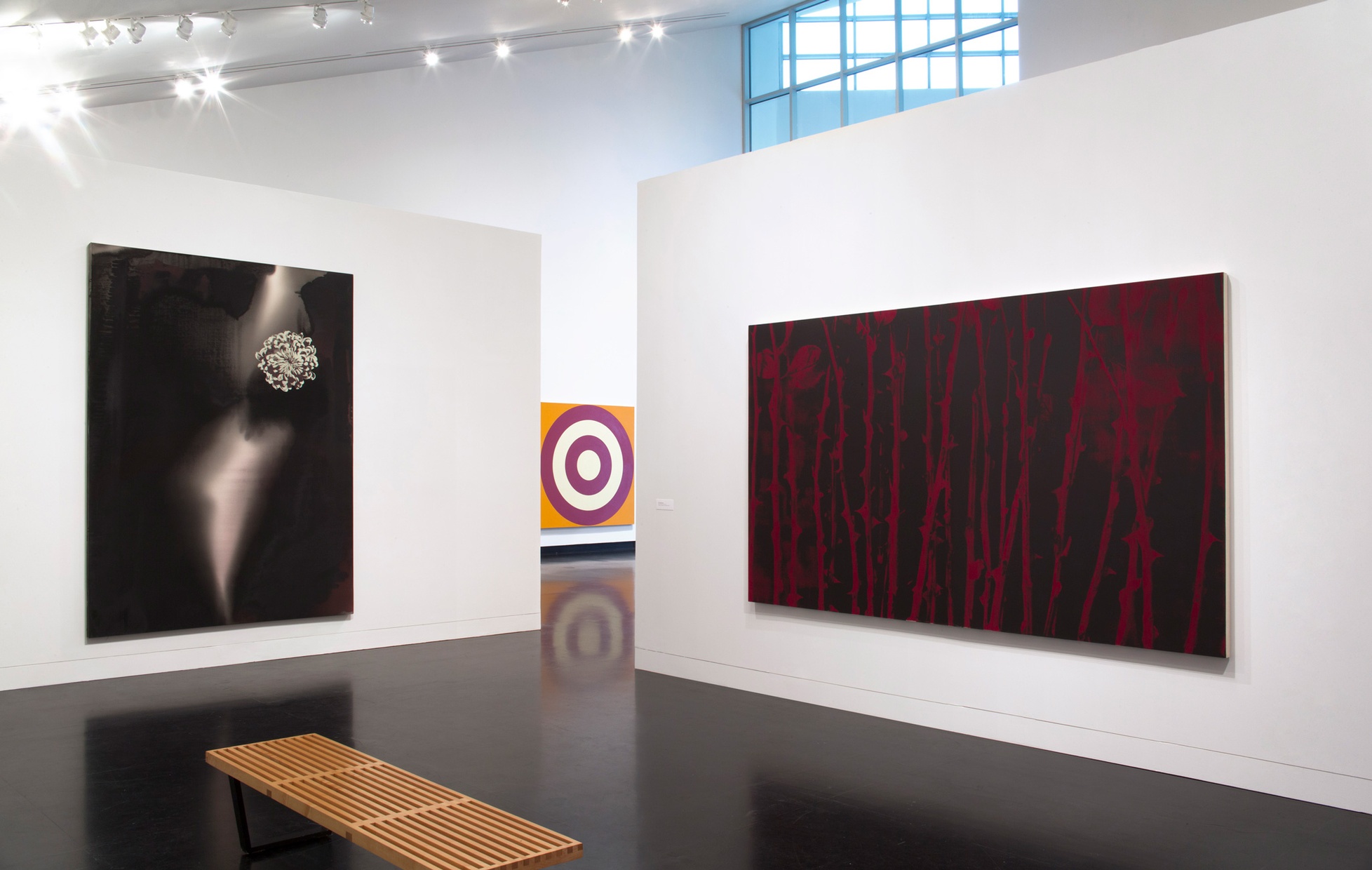 Two large paintings hang on white walls adjacent to each other. On the right, there is a large black painted with red stems and thorns, on the left there is a large black painting with an image of a white flower.