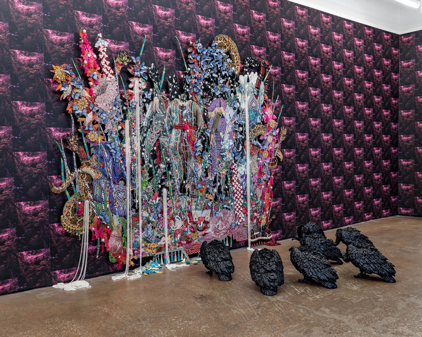 Multimedia installation, featuring a patterned wallpaper on two walls of a gallery with a vibrant, multimedia artwork featuring flowers and beads, with several black bird sculptures on the floor.