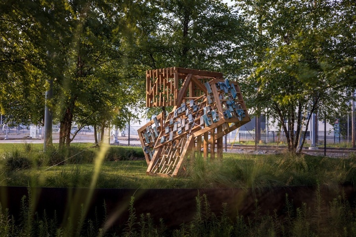 Mixed media large sculpture outdoors in grassy area surrounded by trees with office building in background.