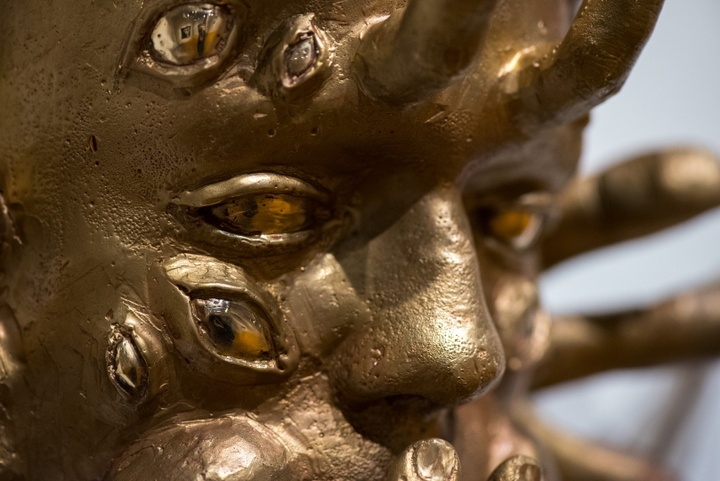 Detail of a bronze sculpture of a face with many amber-colored eyes set into the temples and cheeks. Fingers protrude like horns from the forehead and jawline..