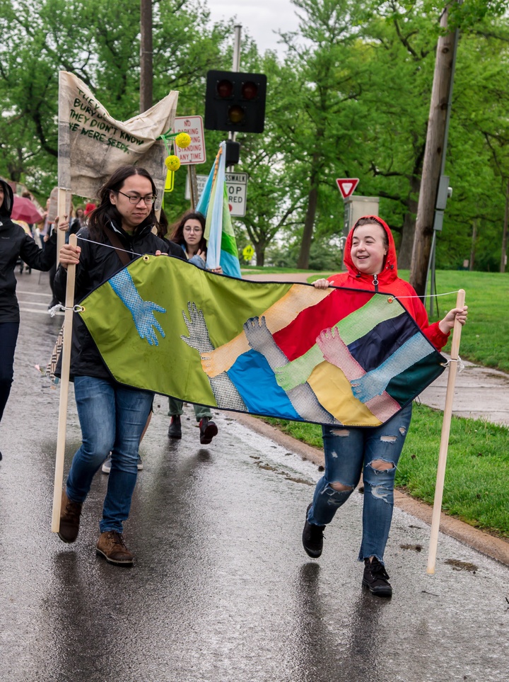 Two people marching in protest holding each ends of the banner which depicts multiple hands holding each other reaching out to a lone hand on the left