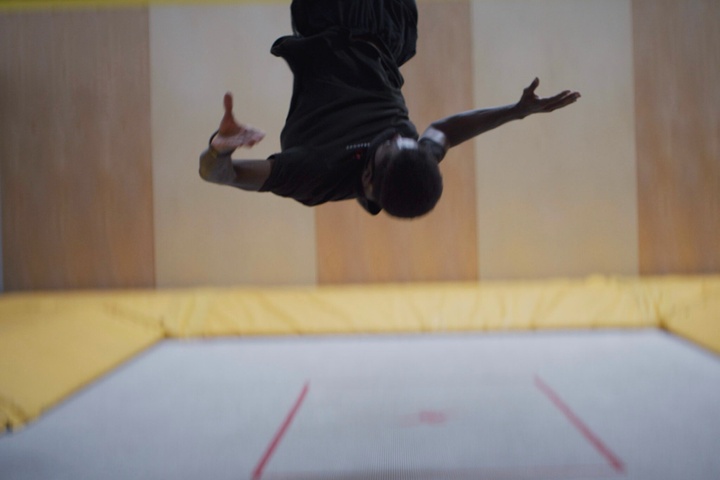 A young man, bouncing indoors on a large trampoline. The camera catches him upside-down, mid-flip, his arms propelling his body around..