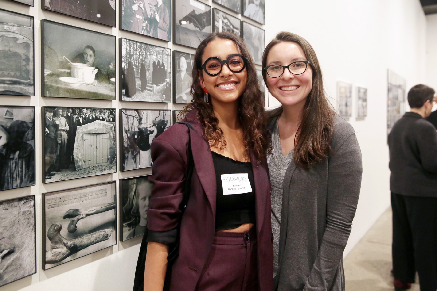 Two young women, one light-skinned and one dark-skinned, stand next to each other smiling in front of a wall with black and white photographs.