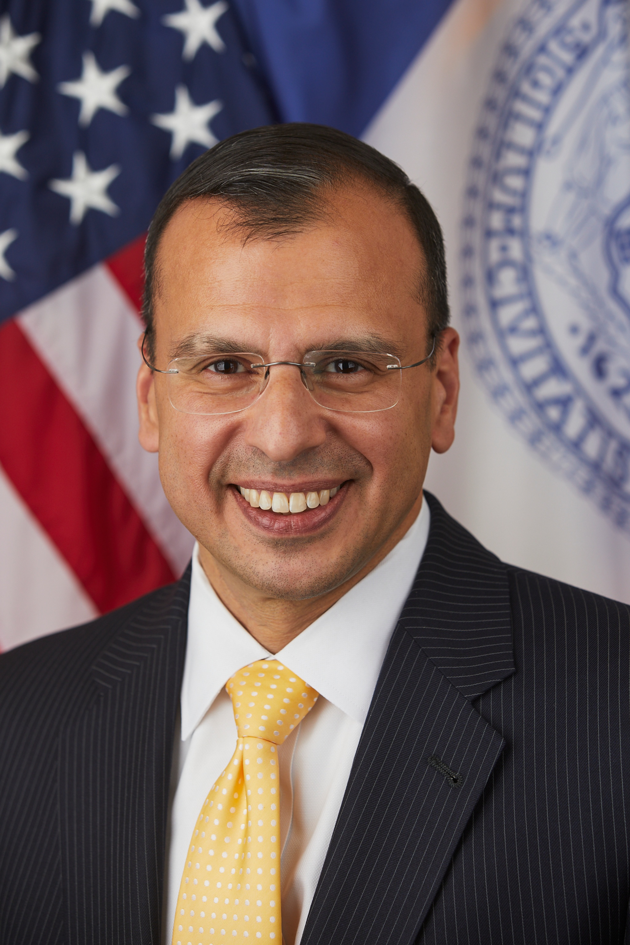 A portrait of Rit Aggarwala. He wears a suit with a yellow tie and smile broadly against a backdrop of the American and NYC flags