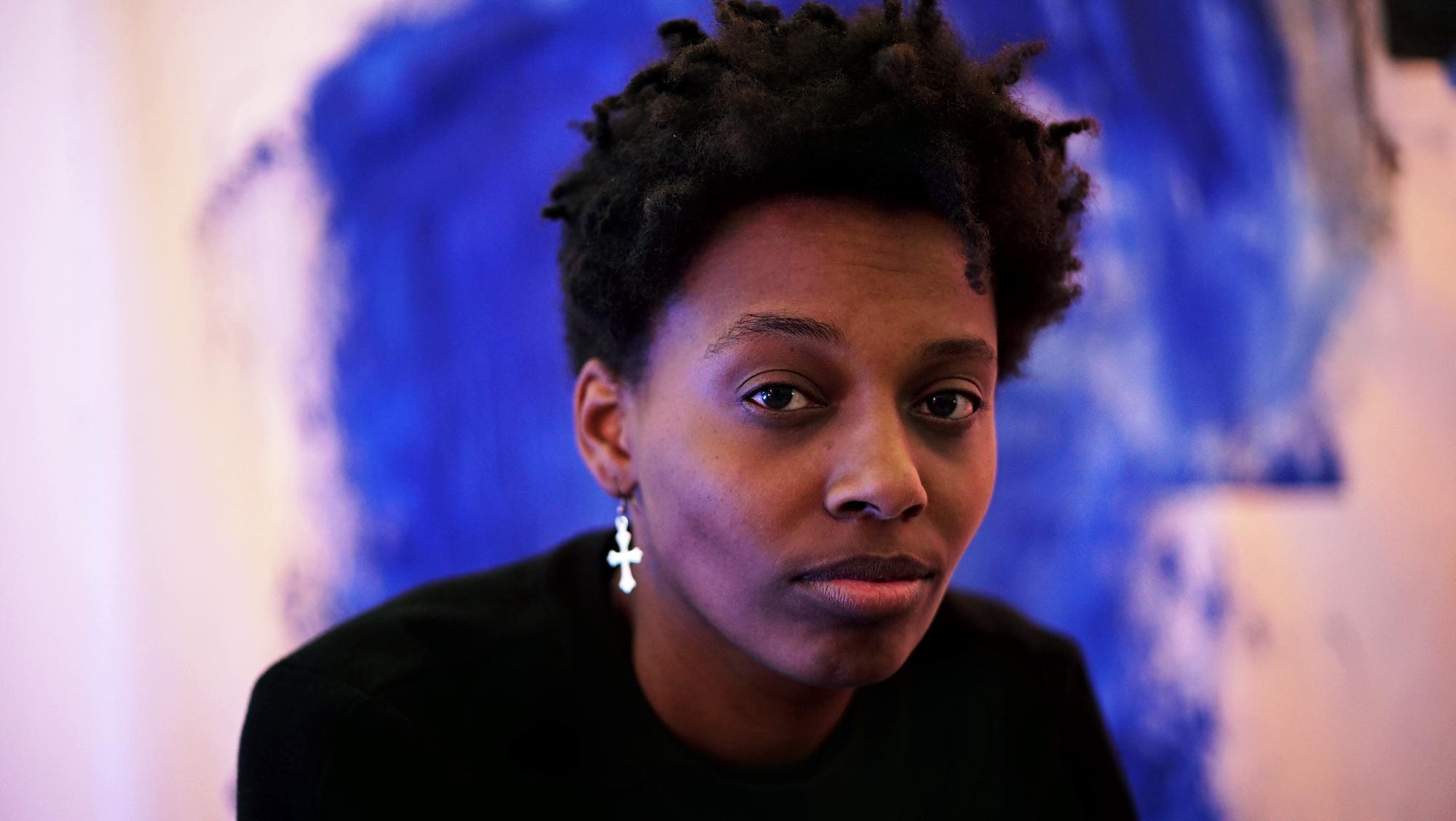A photo of the artist Le'Andra LeSeur in front of a blue painting blurry in the background. LeSeur has short hair and wears a cross earring.