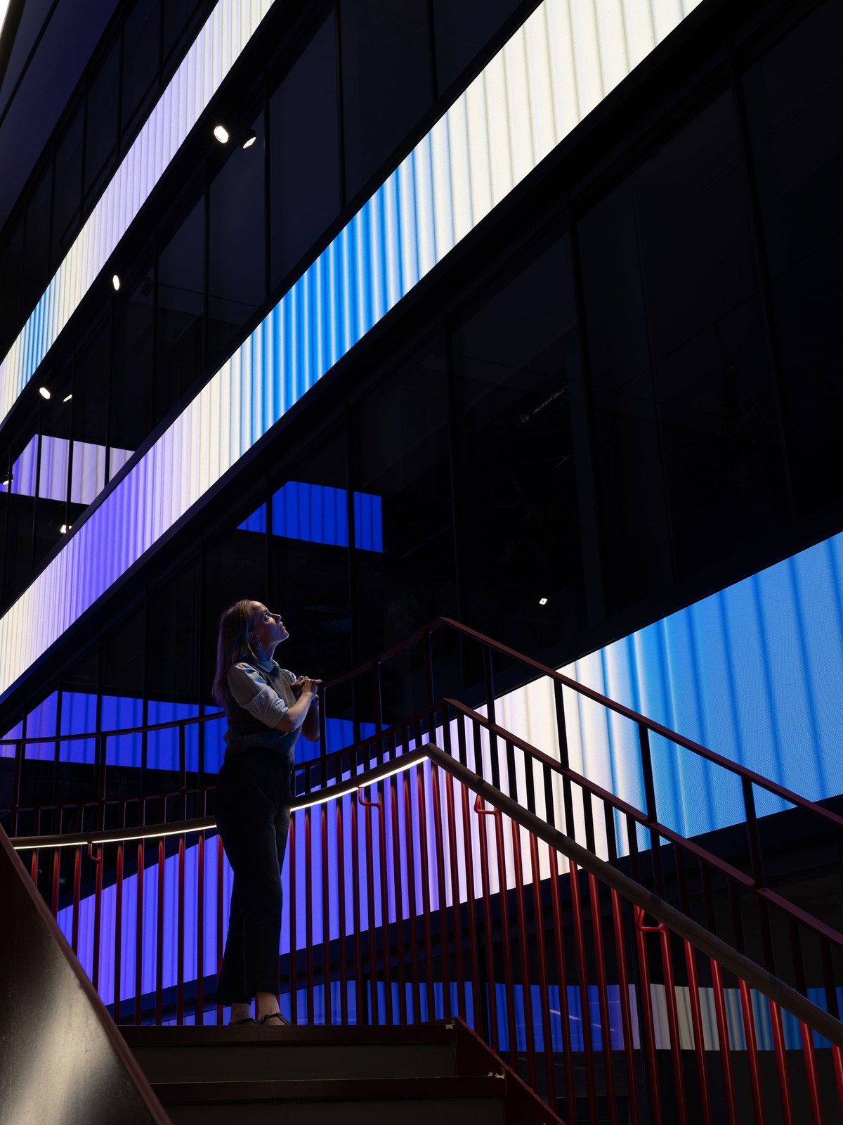 Woman standing on staircase, looking up at installation in atrium, with bands of purple, white and blue