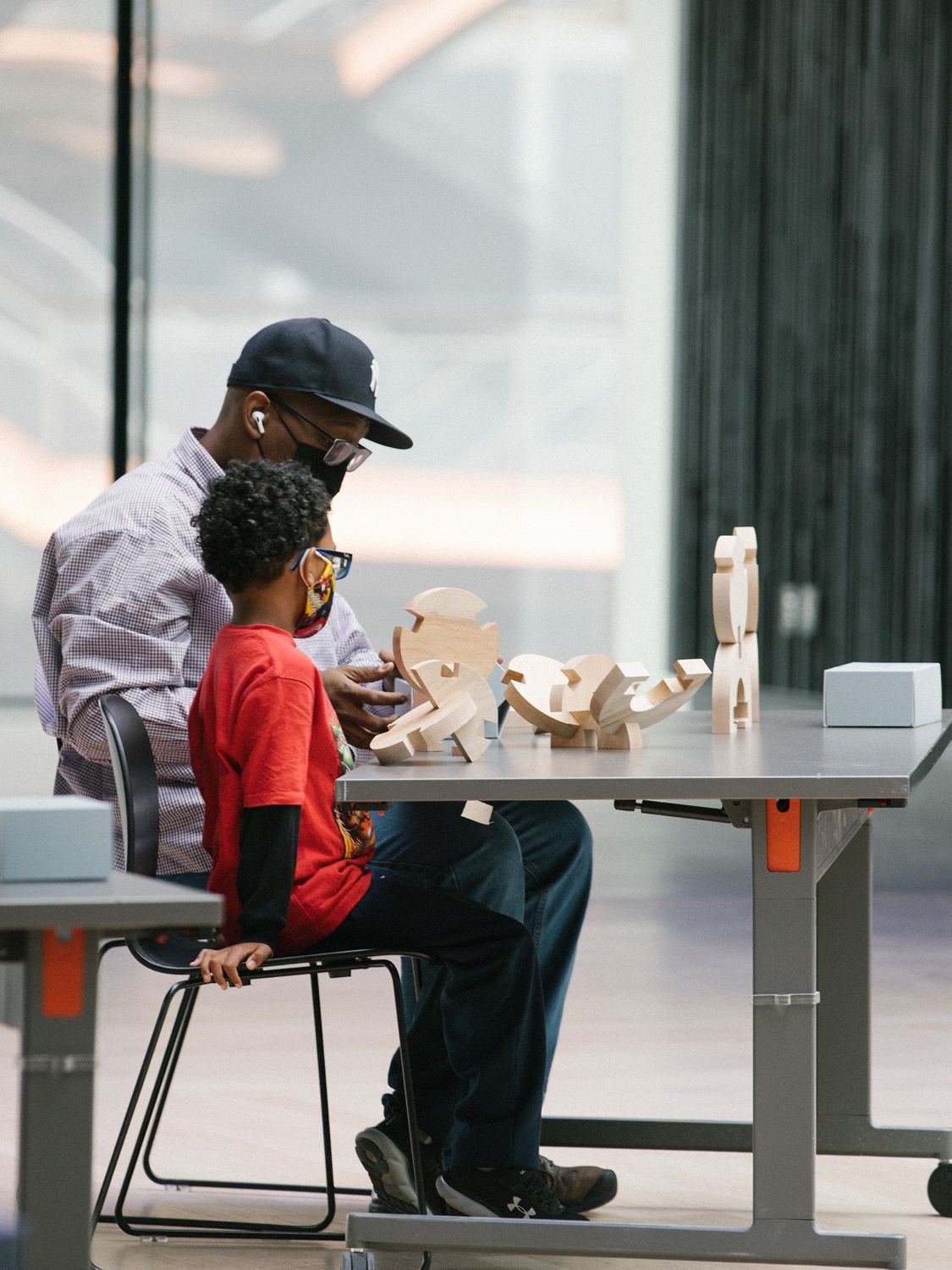 An adult wearing a baseball cap sits at a table with a child in a bright red t-shirt beside him. They are depicted from the side and wooden blocks are on the table.