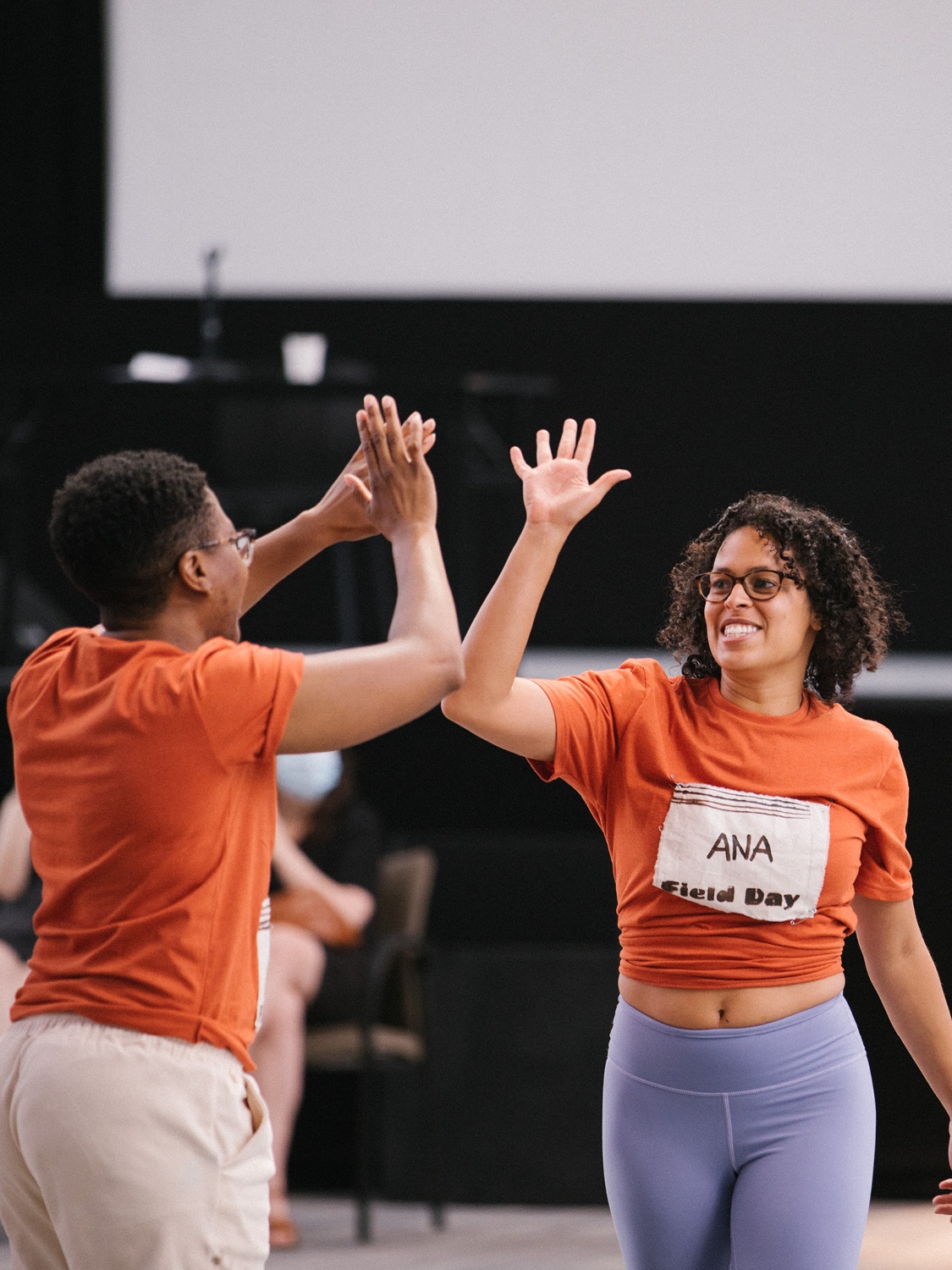 Two people in orange t-shirts lifting their hands to give a high five. The person facing the camera has a name tag across her chest that reads "Ana".