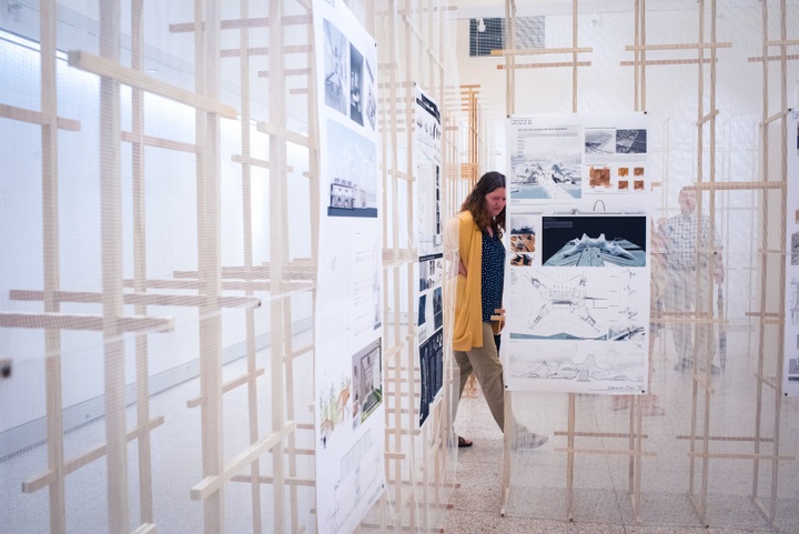 Person walks between display walls constructed of wooden slats and translucent mesh that displays printed architectural rendering images.