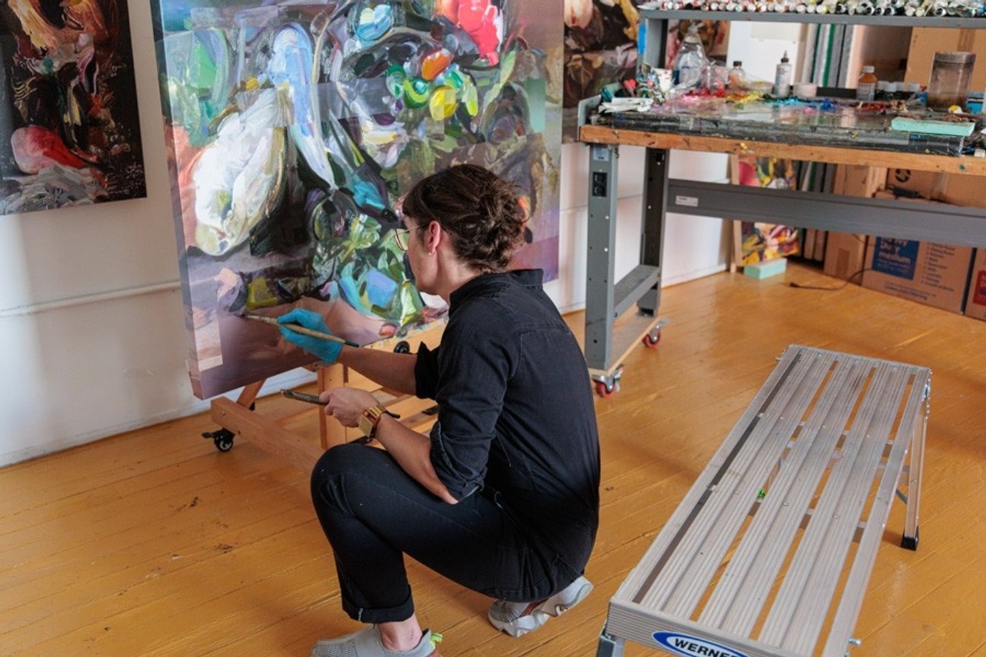 A woman crouches next to a large scale oil painting as she paints. 