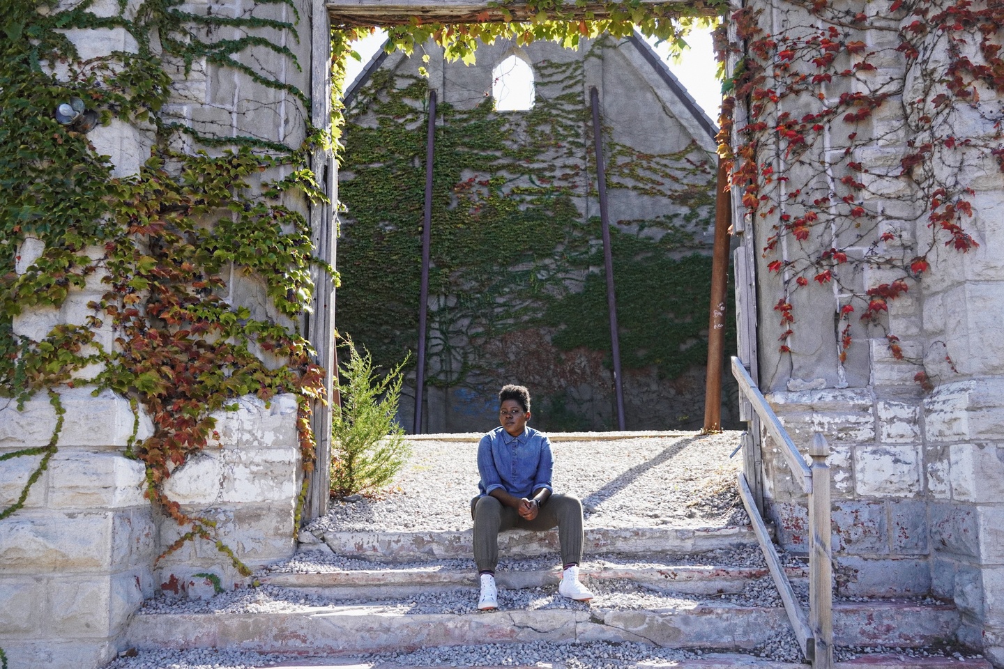 A person sits on the steps of a building with gray walls covered in ivy.