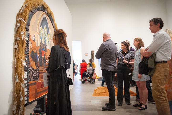 Visitors look at a painting of a Black church sanctuary during a service. The painting is set in an arched frame which is decorated with dry grasses and oyster shells.