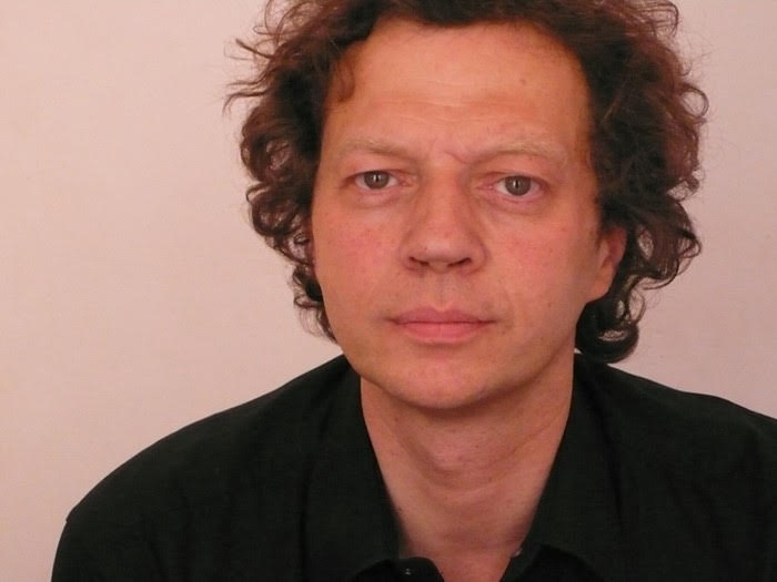 A white man with curly hair looks straight ahead with a quiet intensity. He wears a black shirt and is pictured from the shoulders up against a blank wall.