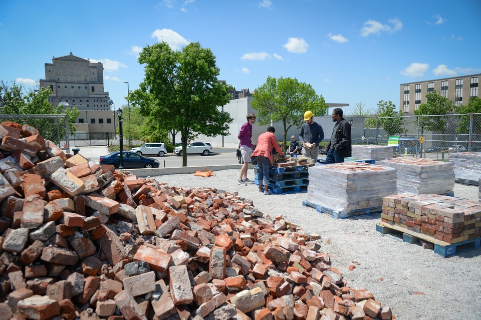 In the foreground, a pile of broken bricks. In the background, a group of people stand around a palette looking a bricks. 