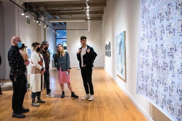 Several people stand and look at works hung on the wall of the gallery. In the foreground is a sheet imprinted with a blue repeating pattern.