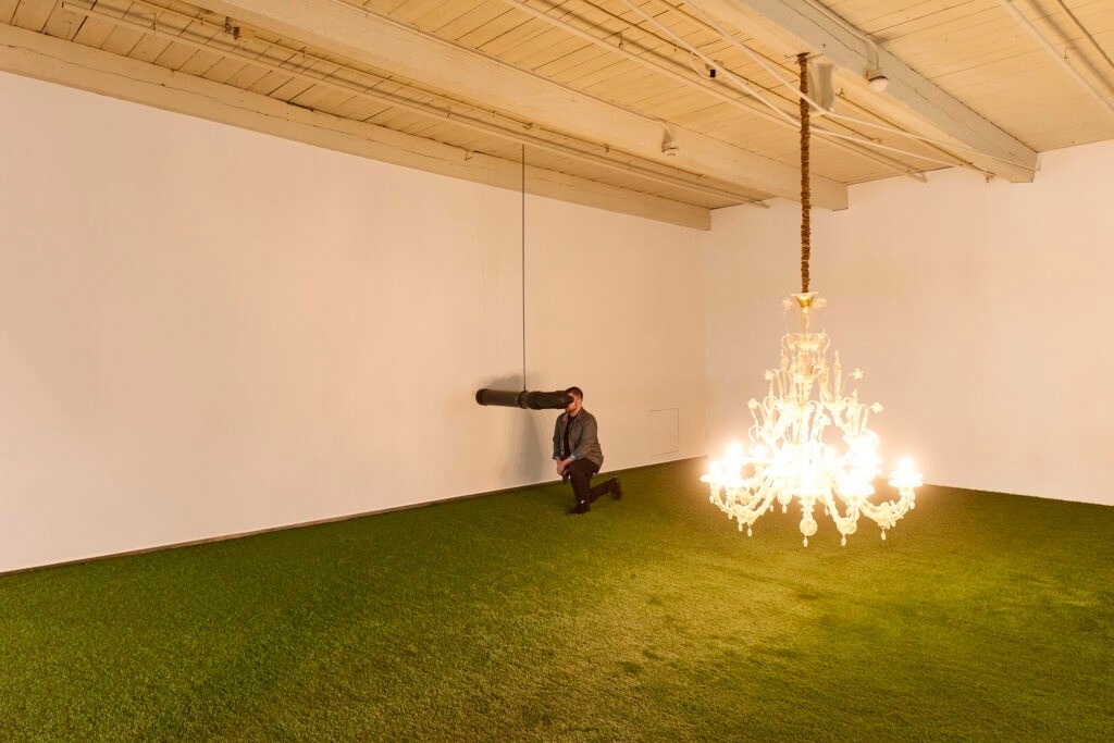 A person peers into a periscope attached to the wall. A chandelier hangs low almost on the ground.