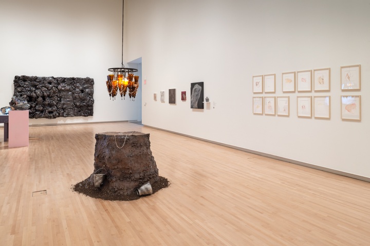 Overview of gallery showing a sculpted tree stump with a chandelier of Aunt Jemima syrup bottles suspended over it, and an array of several light-colored abstract watercolors on the wall.