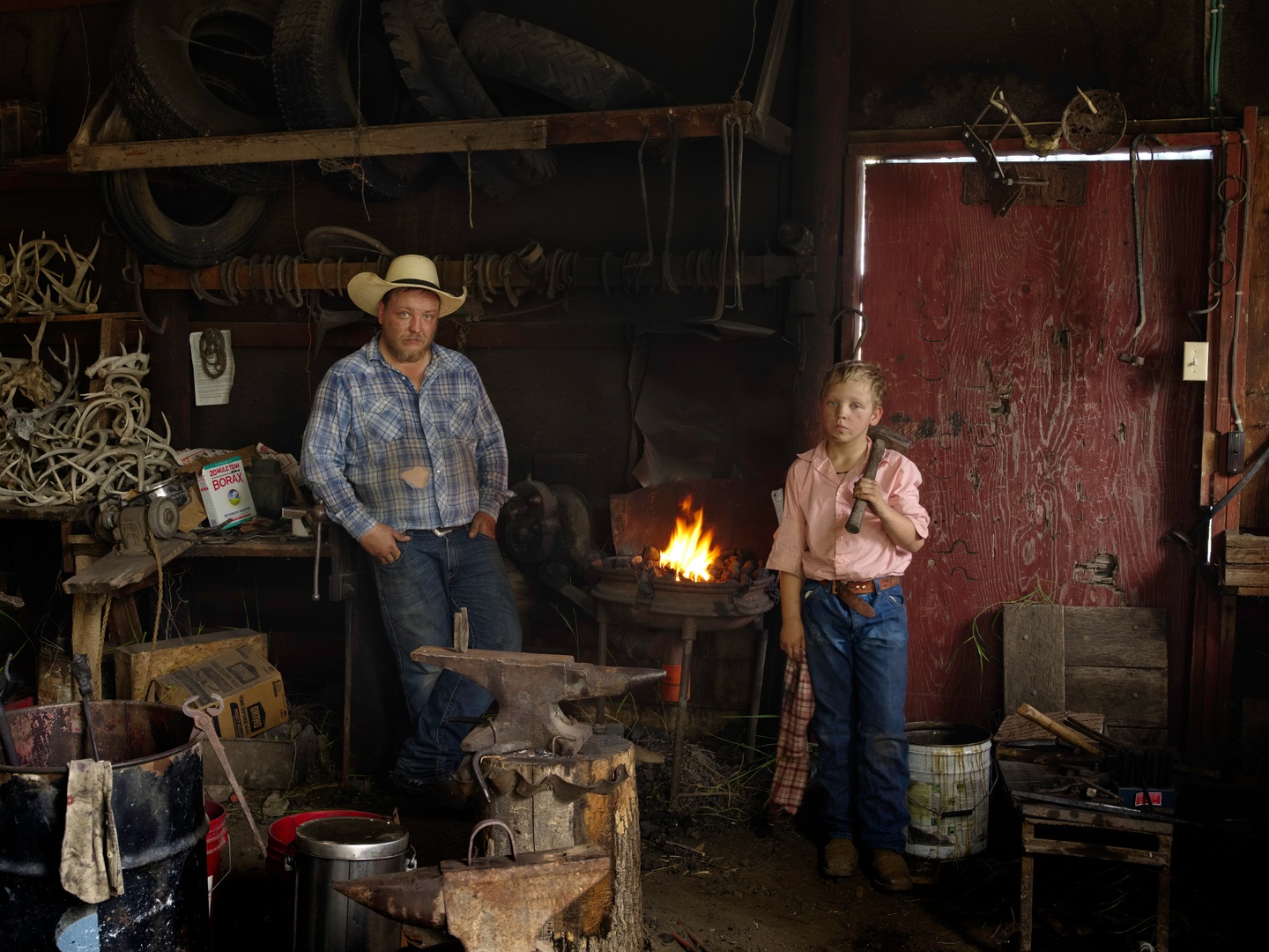 A man and a boy stand in a workshop with a fire behind them and tools