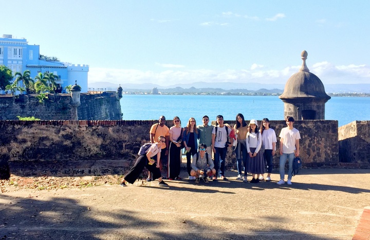 Group of people pose next to an old stone wall built around a city on the waterfront. There are palm trees and a bright blue castle-like structure on another peninsula behind them.