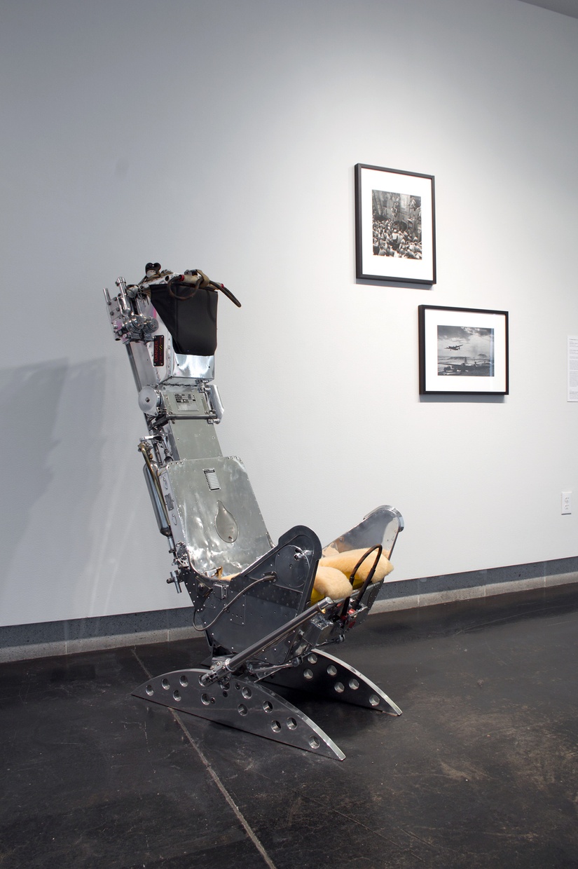 A metal, old-fashioned dentist's chair in a gallery with two framed photographs on the wall next to it.