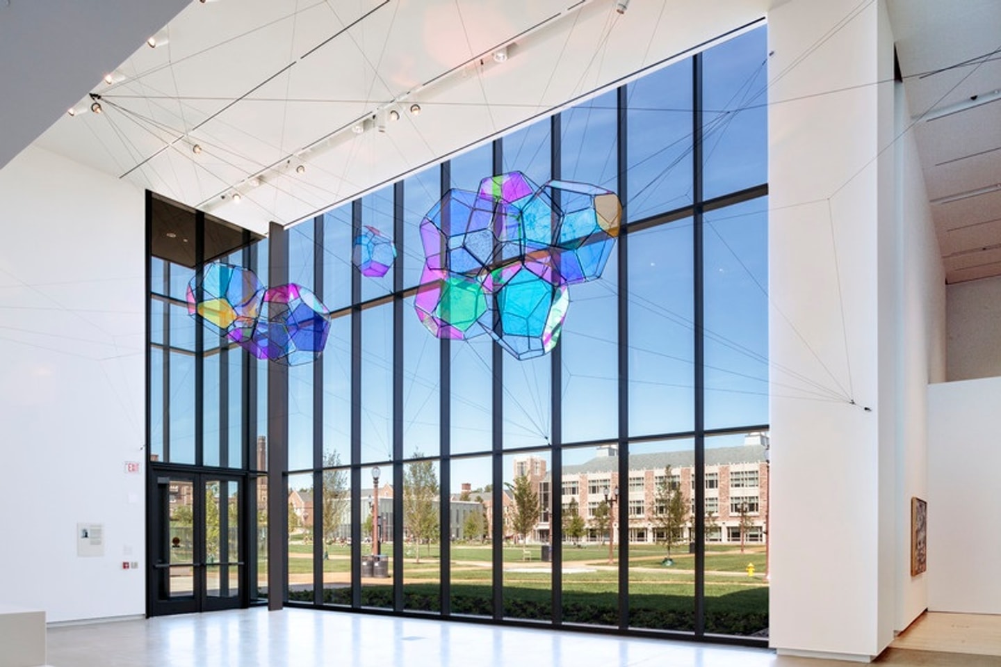 Tomás Saraceno, Cosmic Filaments, 2019. Large clusters of translucent, iridescent geometric forms are suspended from the ceiling by thin black ropes in the museum's atrium. Hanging in front of a wall of tall windows, the installation appears to float, with its colors changing as light filters through the glass.
