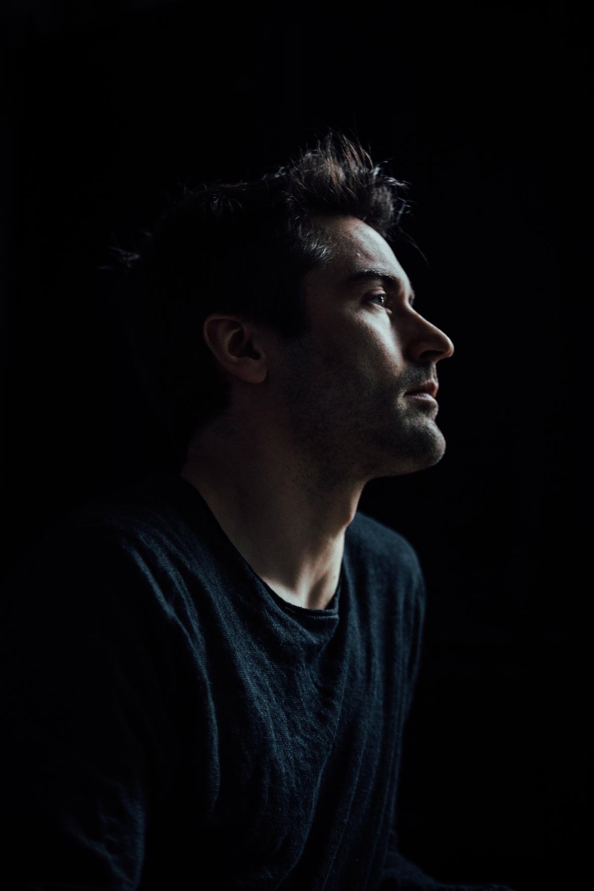 A close-up portrait of pianist Dan Tepfer in a shadowy setting with his face seen in profile in dramatic lighting.