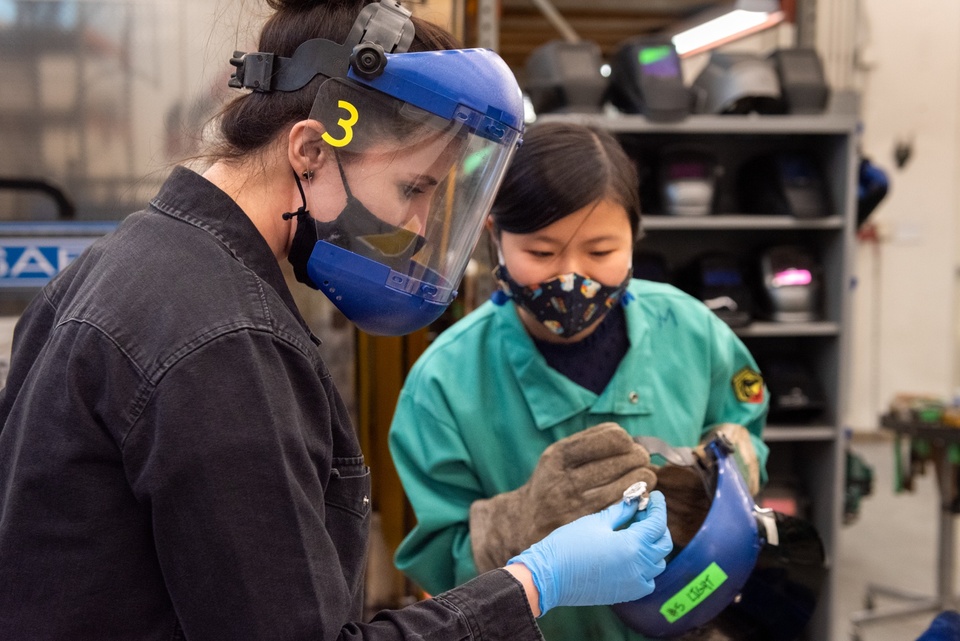 A student and an instructor in face shields and gloves look at a small metal object.