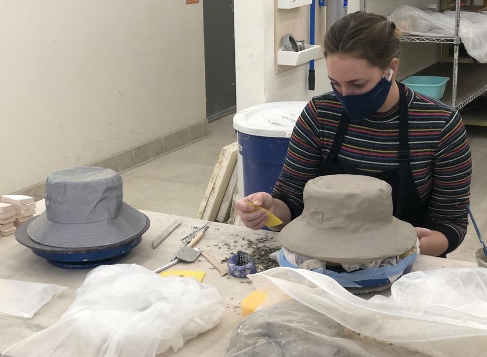 A student works on her clay sculpture of a bucket hat with an actual bucket hat nearby