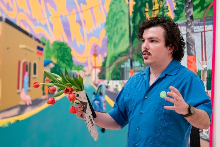 Person holding a bunch of red tulips stands in front of a brightly colored painting of a hallucinatory streetscape and speaks.