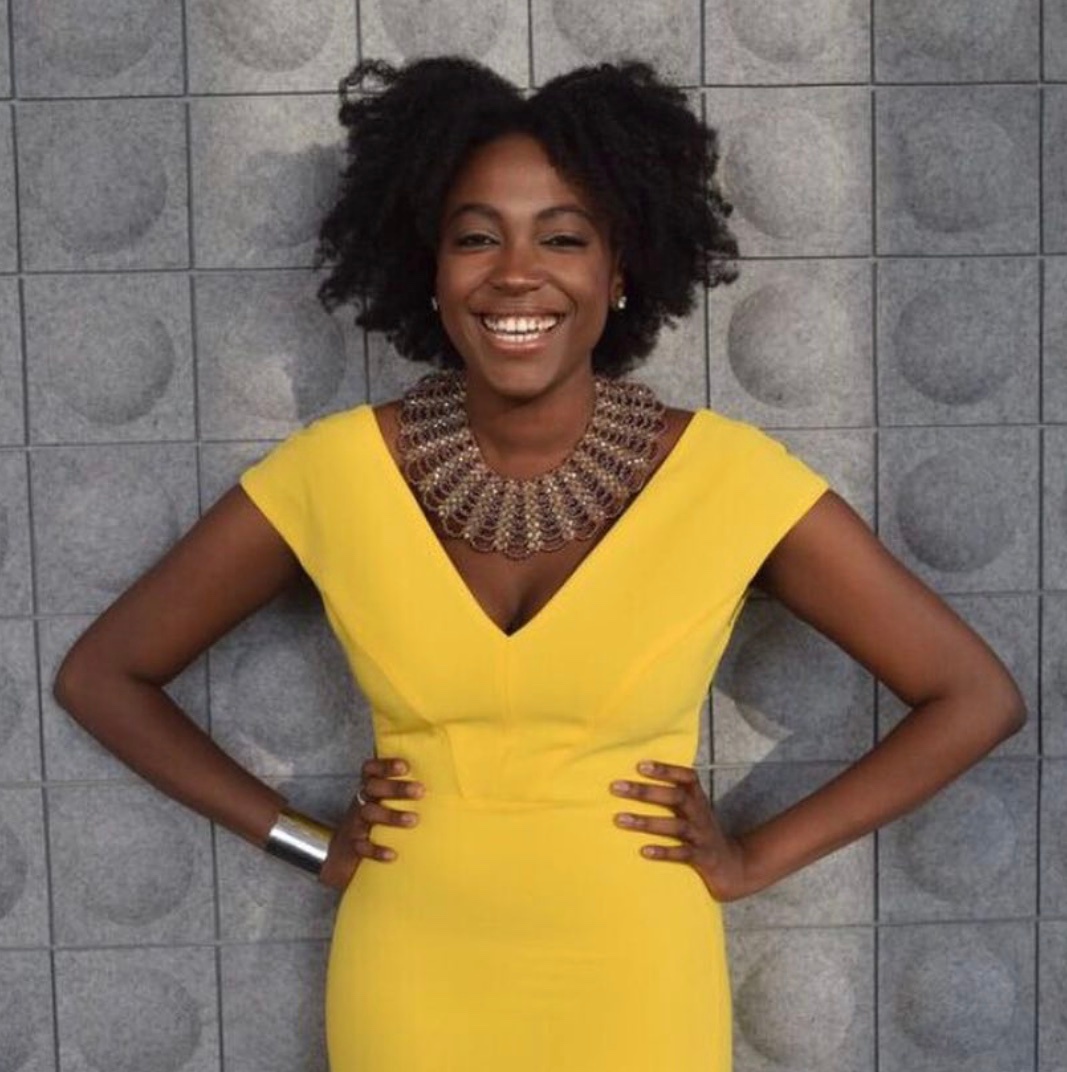 A portrait of Stephanie Quaye, a Black woman with curly shortish hair parted down the center of her head. She wears a bright yellow dress and stands against a gray wall with arms on her hips. She smiles broadly at us. Photo by Imani Schechtman. 