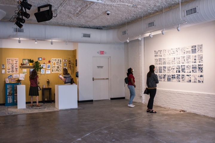 Visitors in a gallery look at a display of comics pages and an installation of a little wallpapered nook with posters, postcards, and pressed flowers fixed to the walls.