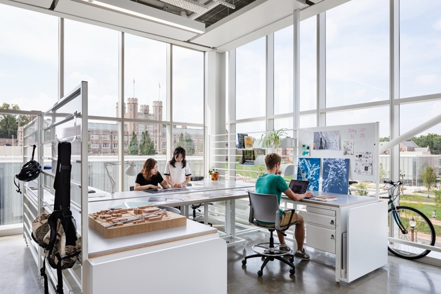 Corner of studio space with several L-shaped desks, floor to ceiling windows, and a view of Brookings Hall.