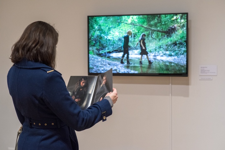 Person flips through a fashion look book in front of a video screen showing a still frame of two children wearing the look book fashions crossing a stream.