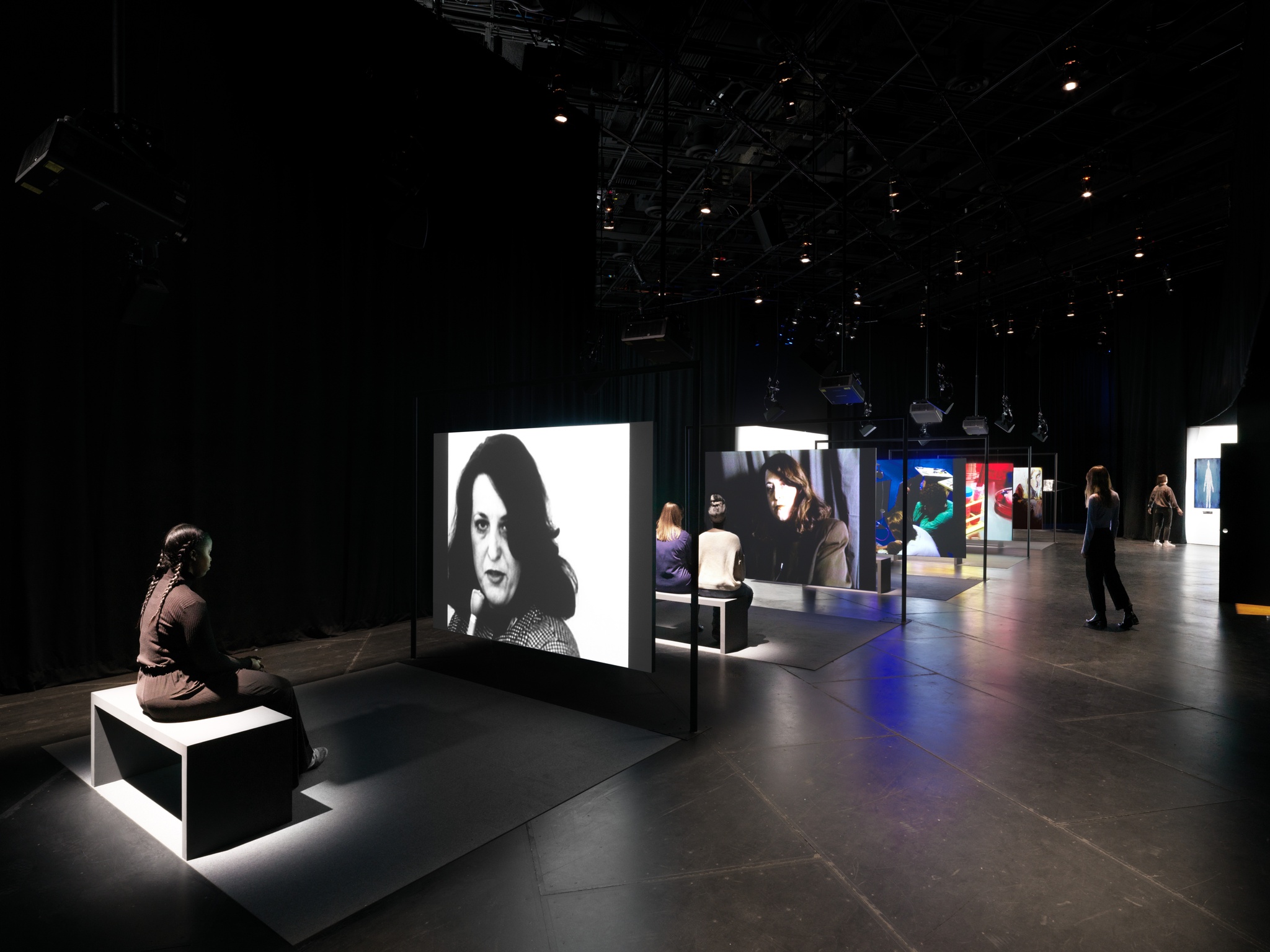 An exhibition visitor sits on a bench watching Lynn Hershman Leeson's Electronic Diaries, with the screens for the multipart work receding into the gallery space